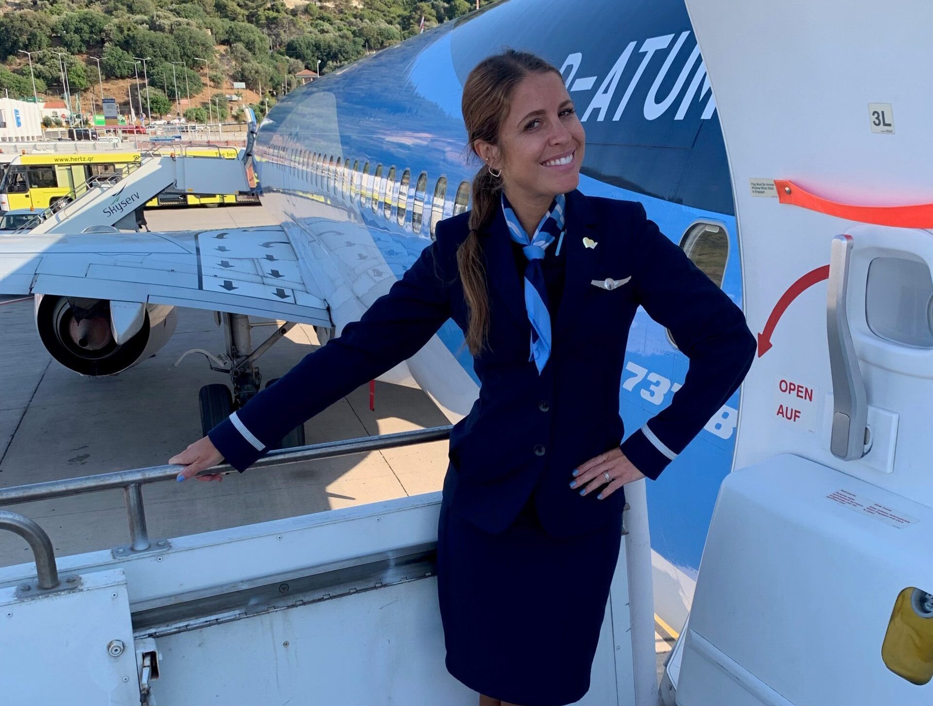 A cabin crew member standing on the steps outside an aircraft. You can see the mountains behind the plane in the background. The cabin crew member is wearing company uniform and smiling.