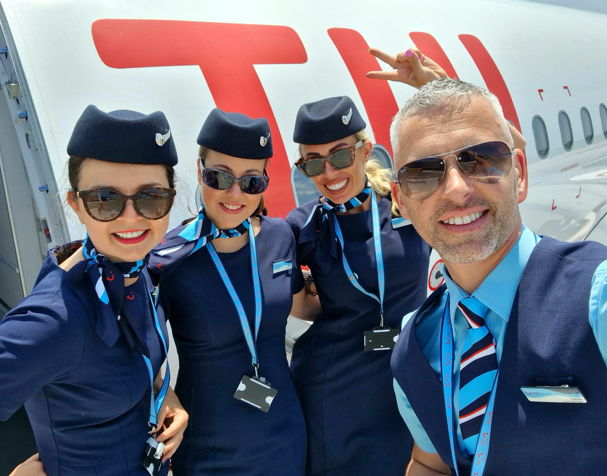 A group of cabin crew members stood on the steps outside the aircraft door. They are smiling with sunglasses on, you can see the TUI text on the plane in the background.