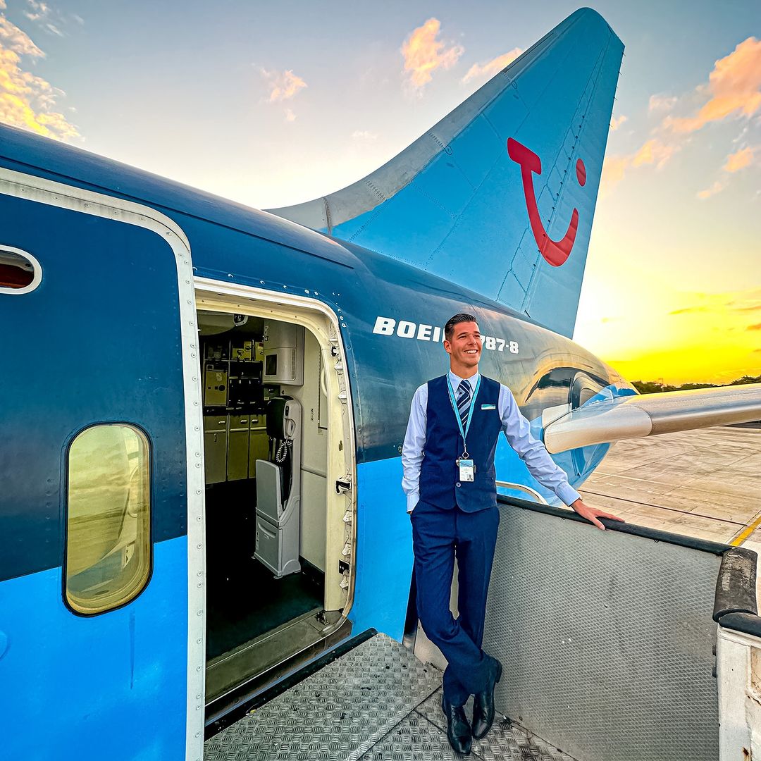 A cabin crew member stood on the ladders outside a plane with a sunset and TUI logo in the background. The cabin crew member is smiling looking into the distance wearing a uniform.