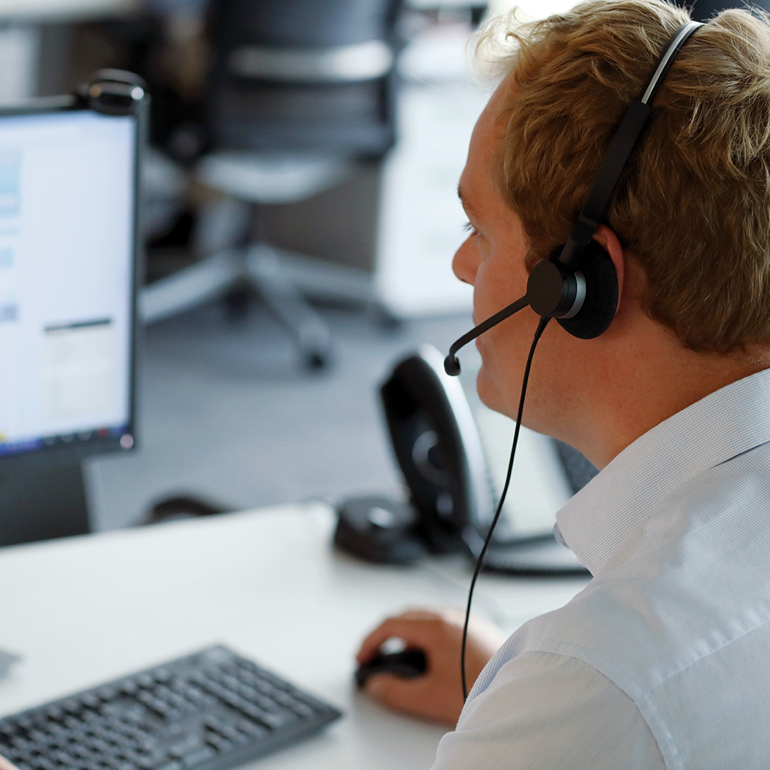 A contact centre agent is looking at their screen with a headset on. There is a mouse and keyboard on the desk.