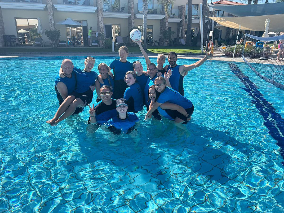 A group of entertainers in a pool having played water volleyboard. They are holding each other up and climbing over each other with guests in the background. They are wearing the TUI Uniform.