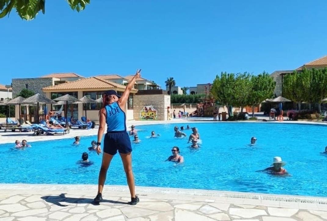 A TUI Entertainer stood in front of a pool which has people in it, their arms are in the air point to the sky. It is a sunnt day, not a cloud in the sky with some green trees in the background on the right, and the hotel on the left.