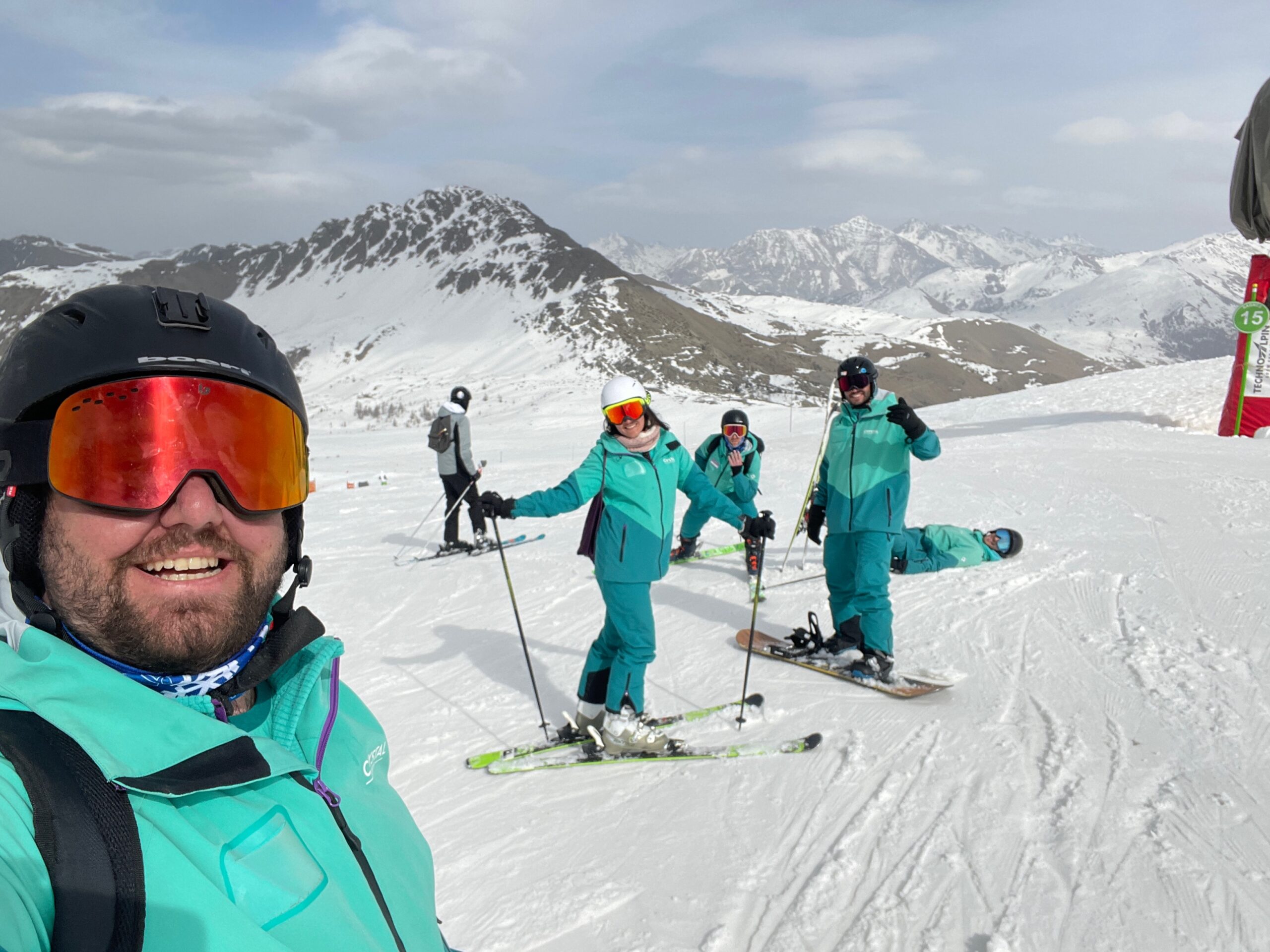 Ski reps on a mountain smiling and laughing. The Ski Reps are stood still wearing goggles and Green Uniforms with the snowy mountain in the background.