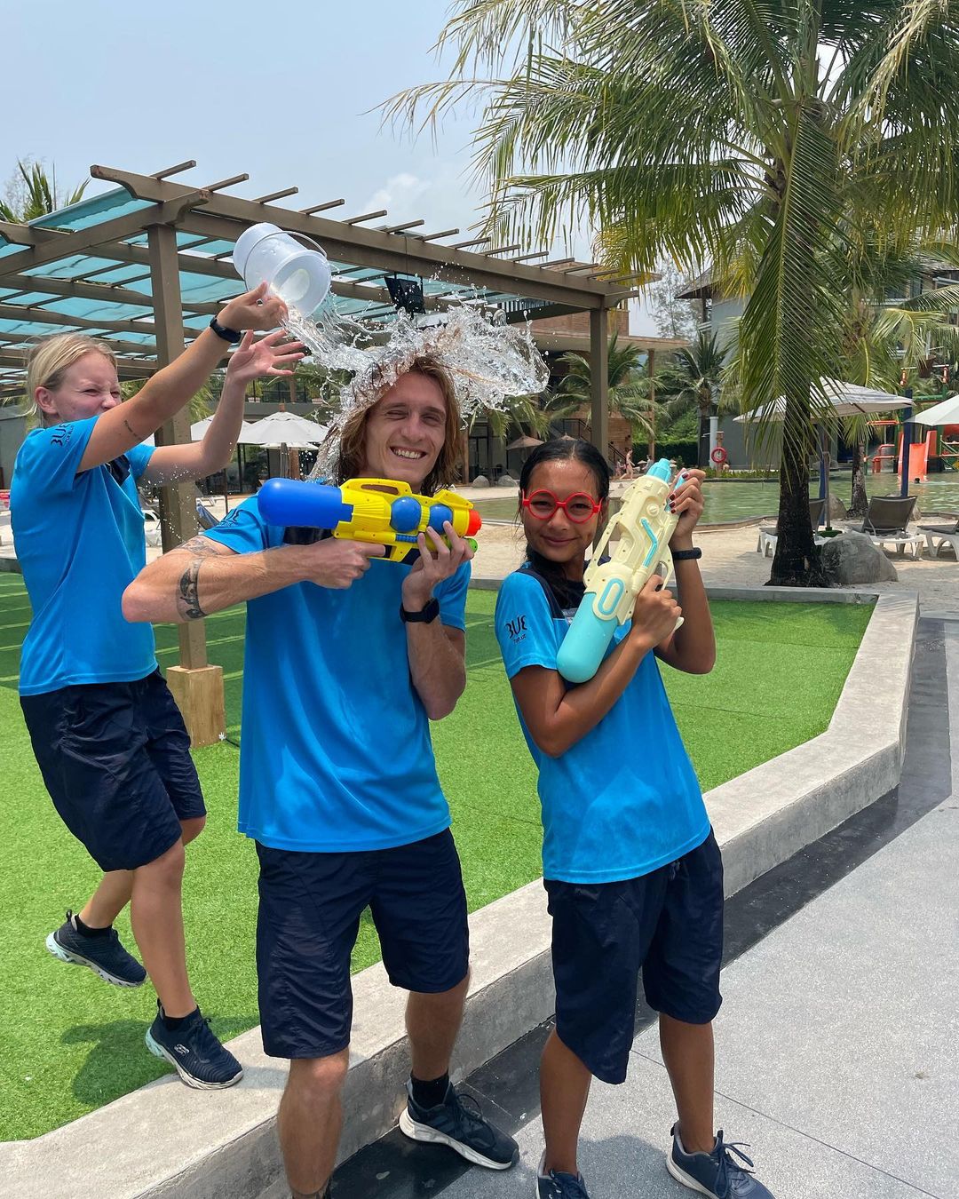 3 TUI entertainers are holding water pistols with one tipping a bucket of water onto someone who has no idea it is happening. They are stood on a pathway with grass behind them. It is a sunny day clearly from the blue sky on show.