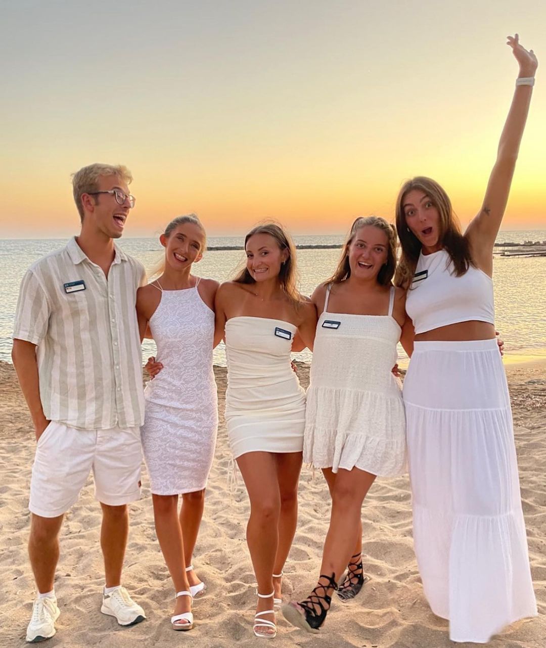 A group of 5 reps are stood on a beach with the sunset behind them. They are smiling with one member of the group having their arm in the air. They are stood on the sand.