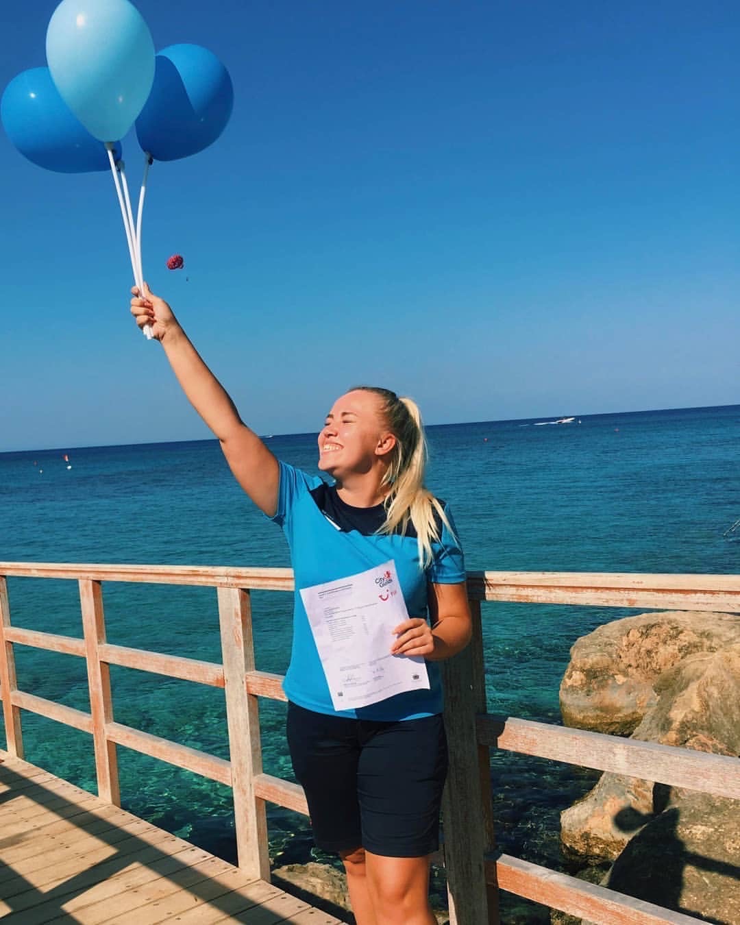 A kids rep working abroad is holding some balloons as she celebrates here childcare qualification. She is wearing a TUI uniform and smiling with the sea and a blue sky in the background. She is stood on a raised promenade.