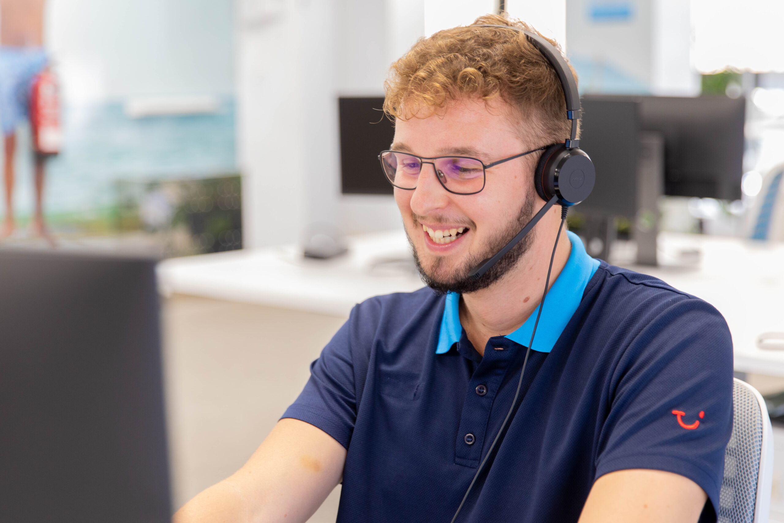 A contact centre employee is smiling at the screen answering calls from a customer. The colleague has glasses and short hair with the office in the background.
