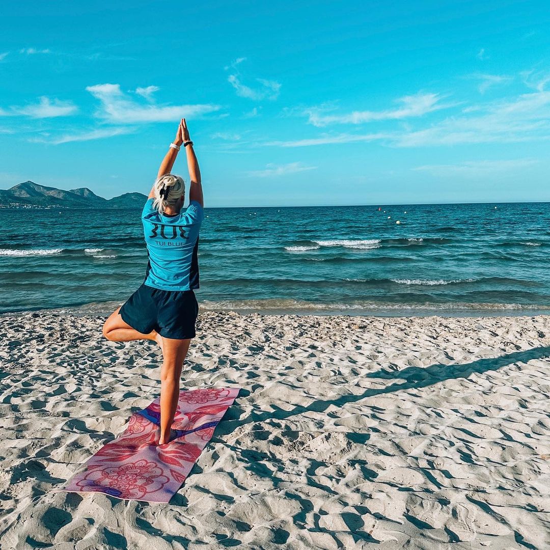 A personal trainer stood doing a Yoga Pose on the beach. There is a towel on the sand underneath where they are stood. You can see white sand, the sea and a blue sky around the personal trainer.
