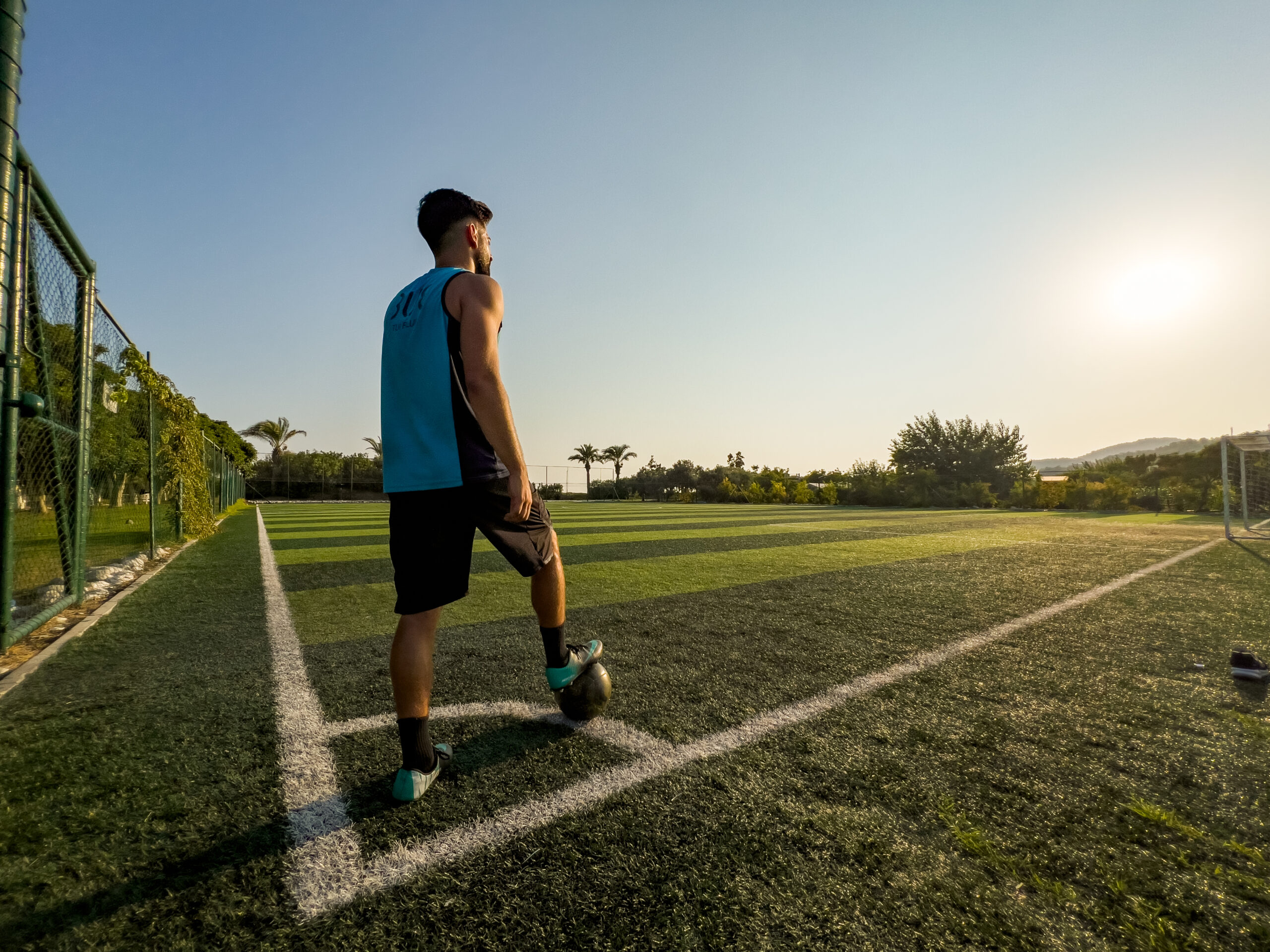 A football coach stood on the corner quadrant of the pitch with a ball under his feet. He is staring out across a football pitch with the sunset clear in view. he is wearing a TUI Football Coach uniform.