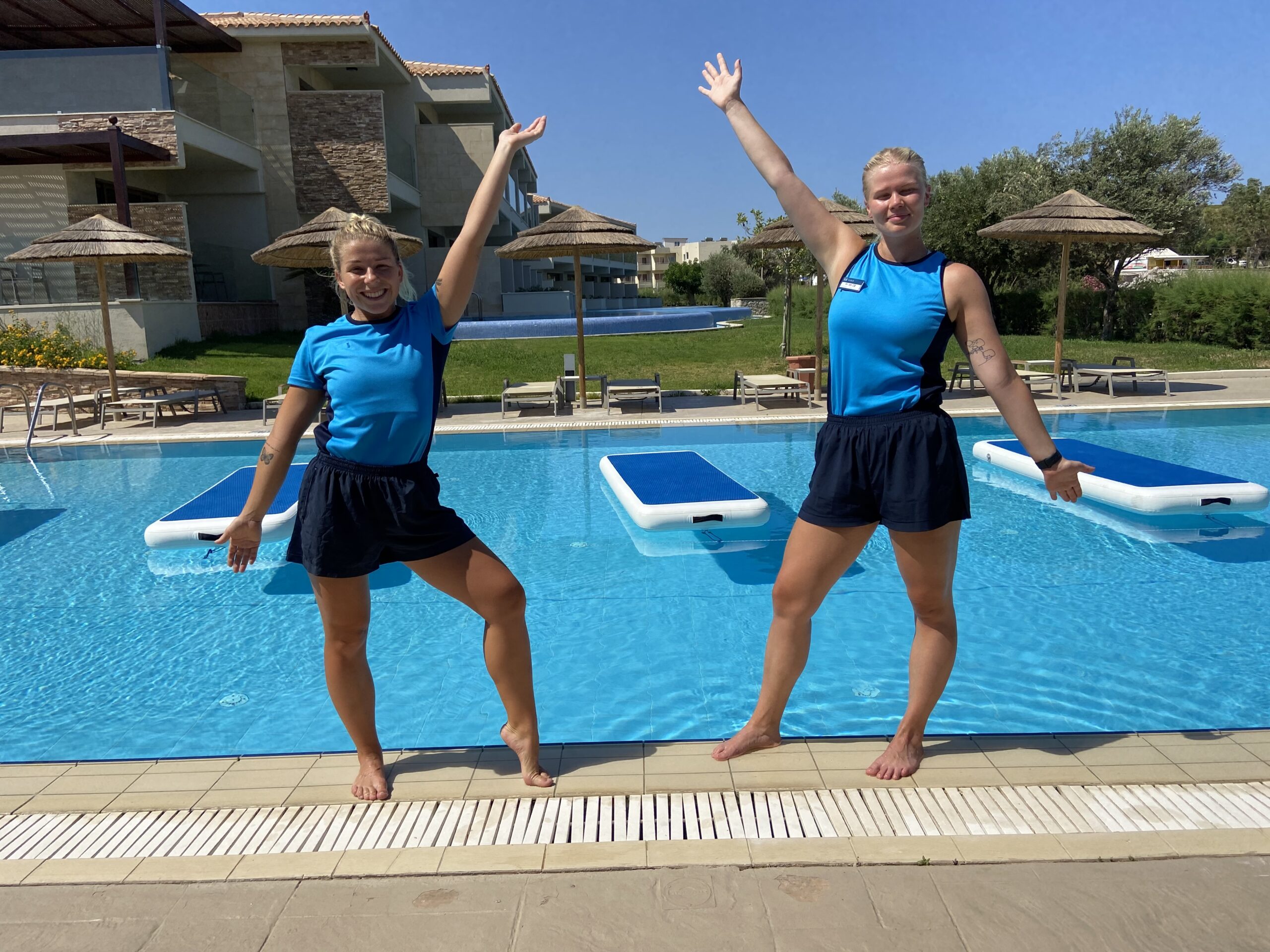 Two swimming instructors stood in front of a pool with their arms in the air smiling at the camera. There is a hotel in the background and it is a sunny day.