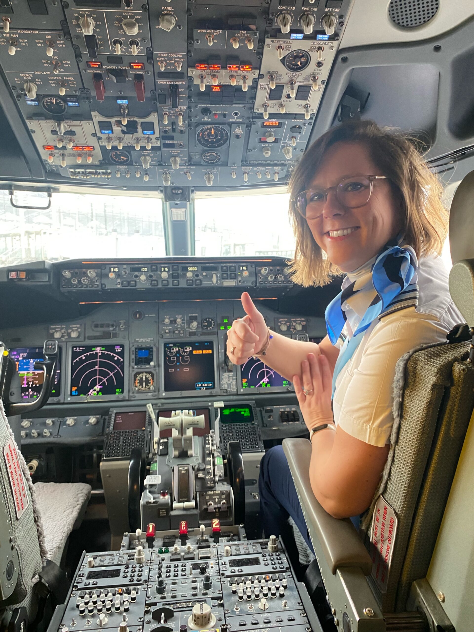 A TUI Pilot sat in the cockpit of an aircraft. The photo is taken towards the cockpit so the pilot is looking back to the camera with the console around and behind them. The pilot is smiling with her thumbs up.
