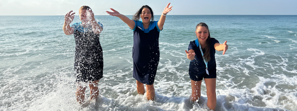 3 reps are stood in the sea, they are playing in the sea with the rep on the left splashing water into the air. They are smiling, wearing the TUI uniform. There is a blue sky in the background.