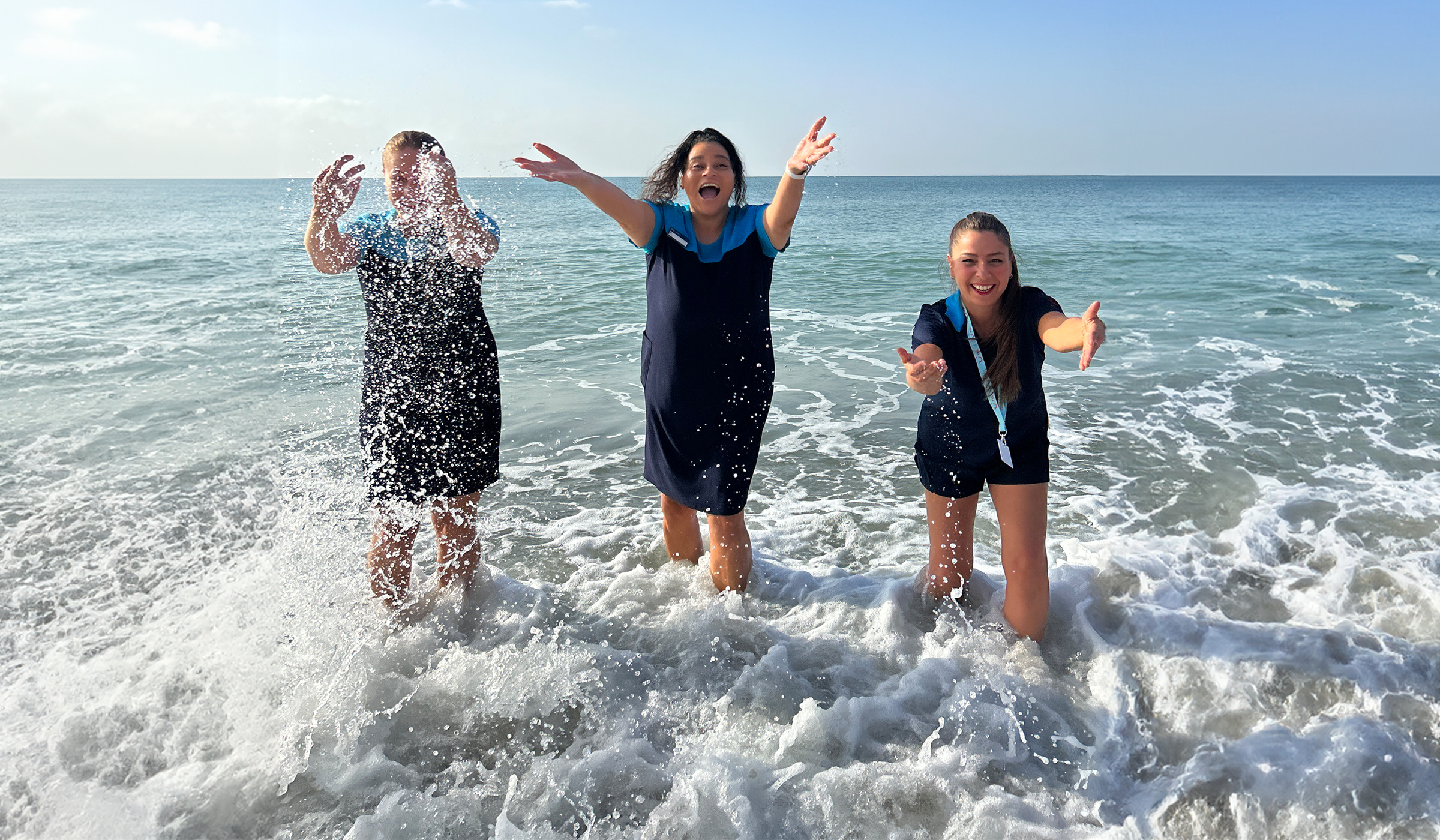 3 reps are stood in the sea, they are playing in the sea with the rep on the left splashing water into the air. They are smiling, wearing the TUI uniform. There is a blue sky in the background.