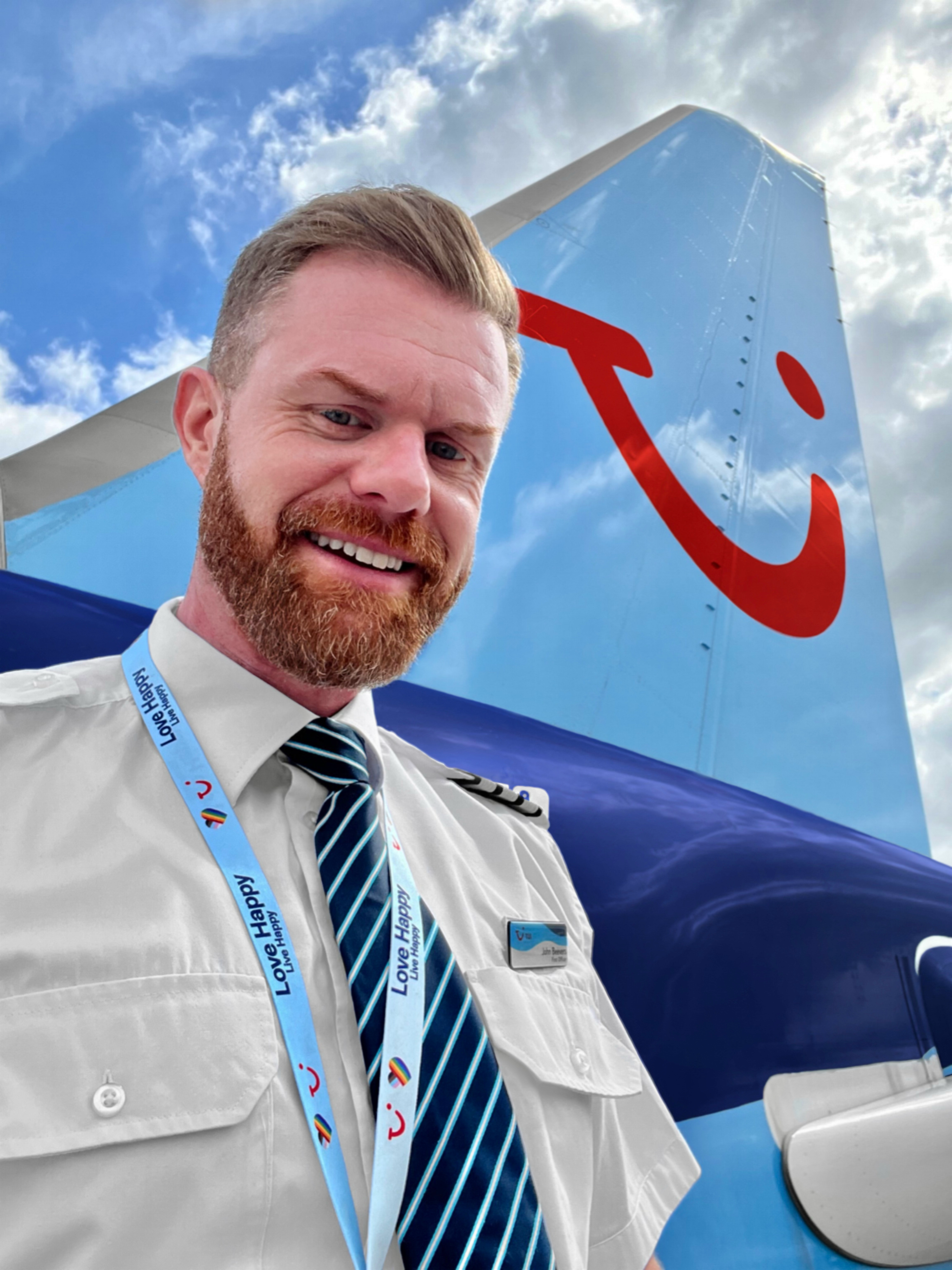 A pilot is stood on the steps of an aircraft. The pilot is wearing their uniform with the fin of the plane behind him. There is a blue sky with a few clouds.